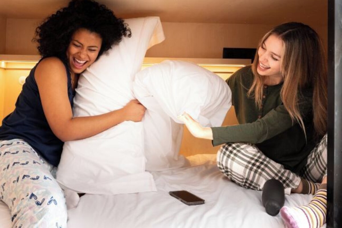 Two people sitting on a bed with pillows, a smartphone on the bed, and a wall-mounted light fixture above.