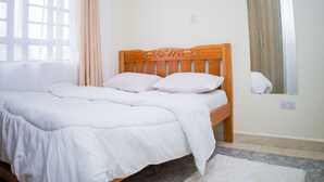 Una cama bien hecha con ropa blanca y un cabecero de madera, una ventana con cortinas de gasa y un espejo en la pared.