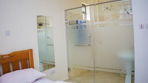 Un dormitorio con cabecera de madera, cama con ropa de cama blanca, cabina de ducha de vidrio y baño con inodoro y lavamanos.