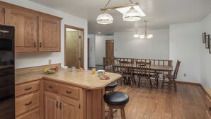 Uma cozinha com armários de madeira, uma ilha central com banquetas, uma mesa de jantar com cadeiras e iluminação pendente.