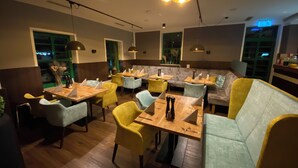 A restaurant with wooden tables and chairs, a green window, and a blue light on the ceiling. - Hotel & Restaurant Elbschetal (Wetter)
