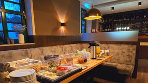 A breakfast buffet setup with a variety of food items, a coffee station, and a seating area with a sofa and a wooden table. - Hotel & Restaurant Elbschetal (Wetter)