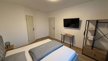Een slaapkamer met een bed, een aan de muur bevestigde televisie, een houten garderobe en een kleine tafel met een lamp.