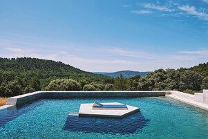 Una piscina con un área de descanso flotante, rodeada de exuberante vegetación y colinas al fondo.