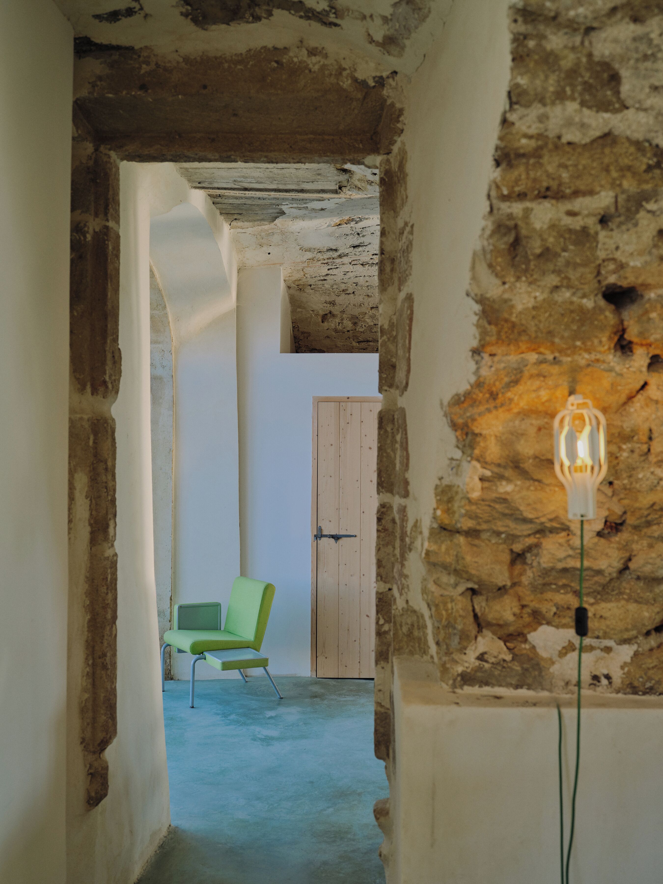 A narrow corridor with exposed brick walls, a wooden door, and a green chair.