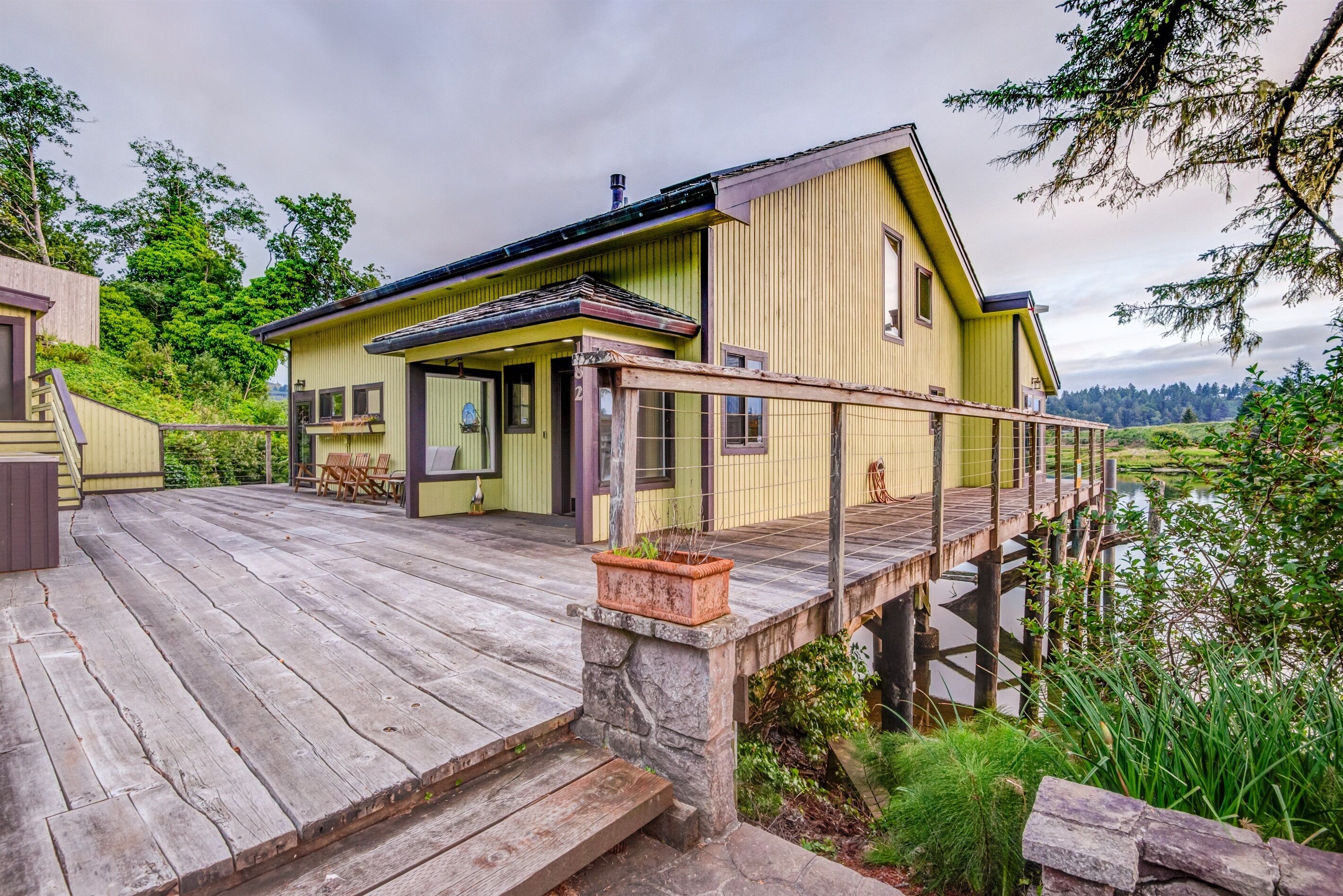 A wooden deck leading to a yellow building with a stone base, overlooking a body of water.