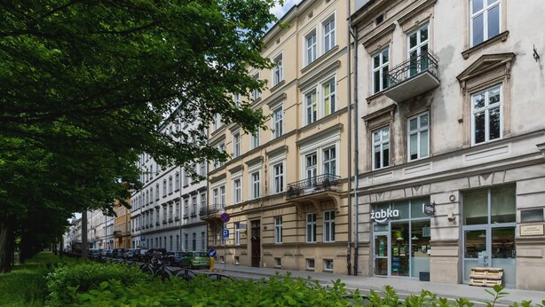 A row of multi-story buildings with balconies, a storefront with a sign, and parked cars along a tree-lined street. - Comfortable Apartment in the Center | Close to Wawel Castle | Smart TV (Kraków)