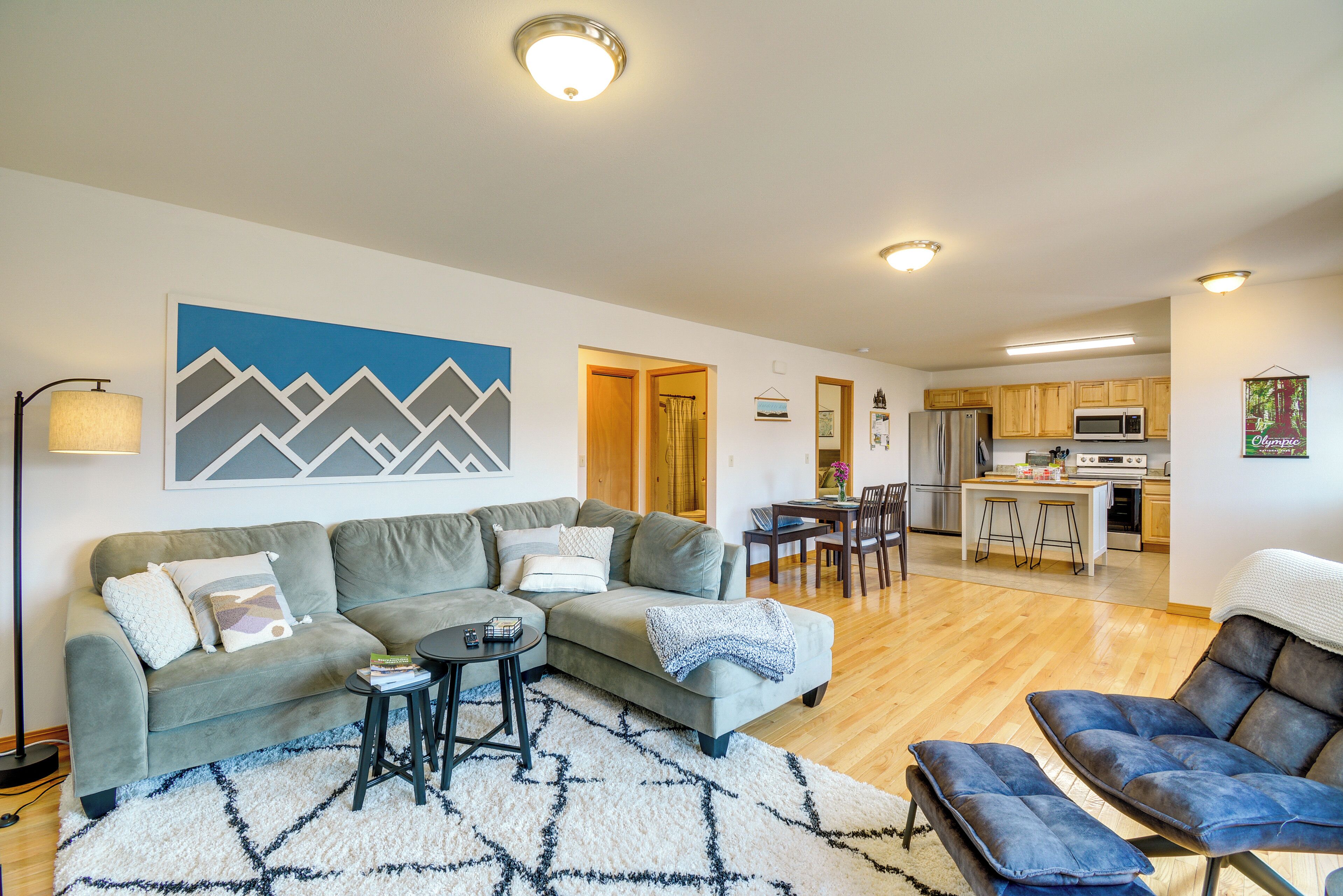 A modern living room with a sofa, coffee table, and ottoman, a dining area with a table and chairs, and a kitchen with appliances.