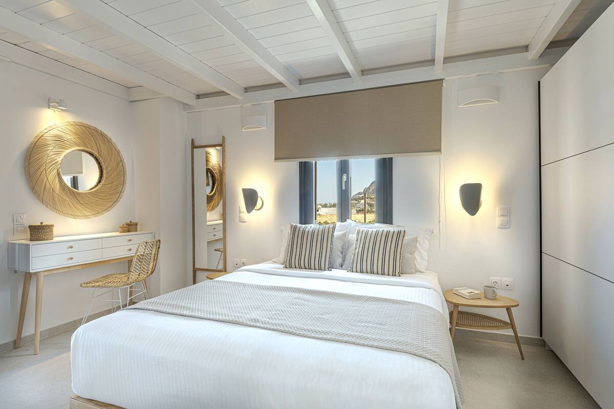 Une chambre avec un grand lit, un bureau, une chaise, un miroir rond et une vue sur l’extérieur.