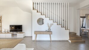 A modern living room with a staircase, a flat-screen TV, a console table, and a dining area. - Escape View Villa (Santorini)