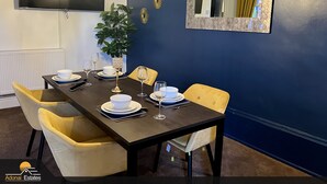 Uma área de refeições com parede azul escura, uma televisão de tela plana grande, espelhos decorativos e um vaso de planta.