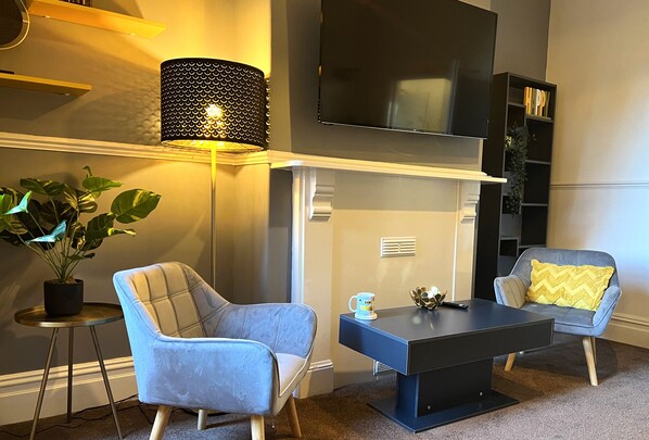 Un salon moderne comprenant un téléviseur à écran plat, deux fauteuils, une table basse, une lampe sur pied et une bibliothèque.