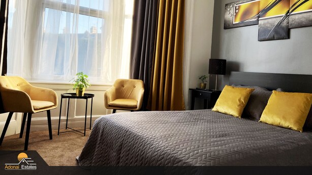 Quarto de hotel com uma cama grande, duas poltronas, uma pequena mesa com uma planta e uma mesa de cabeceira com abajur.