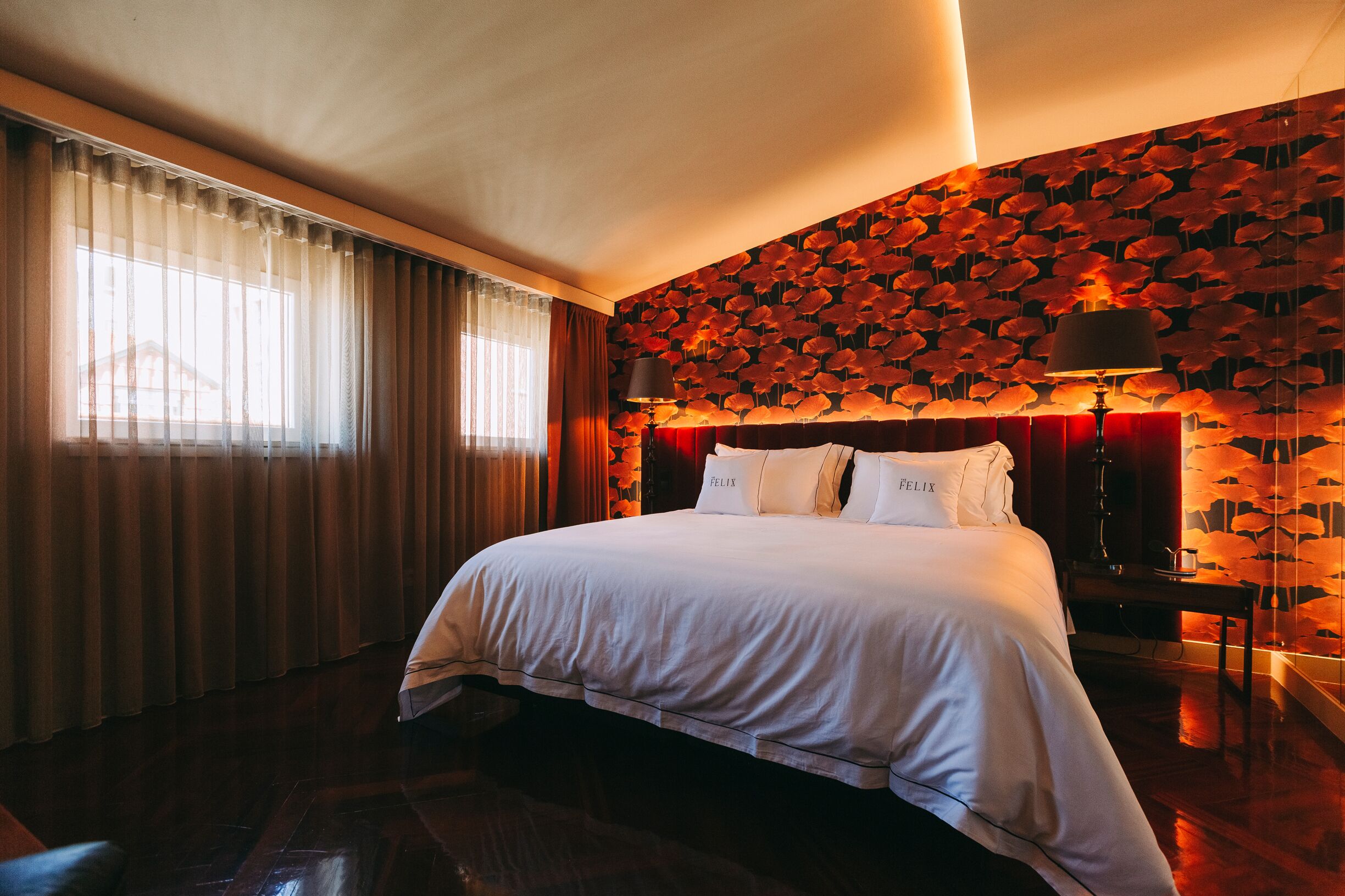 A hotel room with a large bed, a wall with a leaf pattern, a window with curtains, and a nightstand with a lamp.