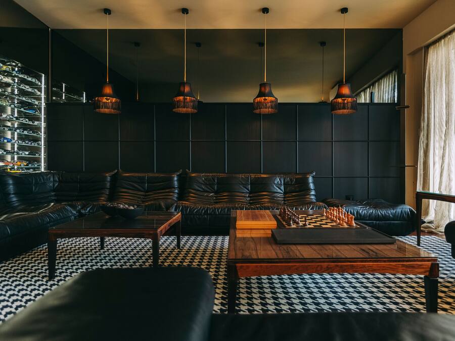 Un salon moderne avec un canapé en cuir noir, une table basse en bois et un jeu d’échecs. Des lampes suspendues illuminent l’espace, et le sol est recouvert d’un motif en noir et blanc.