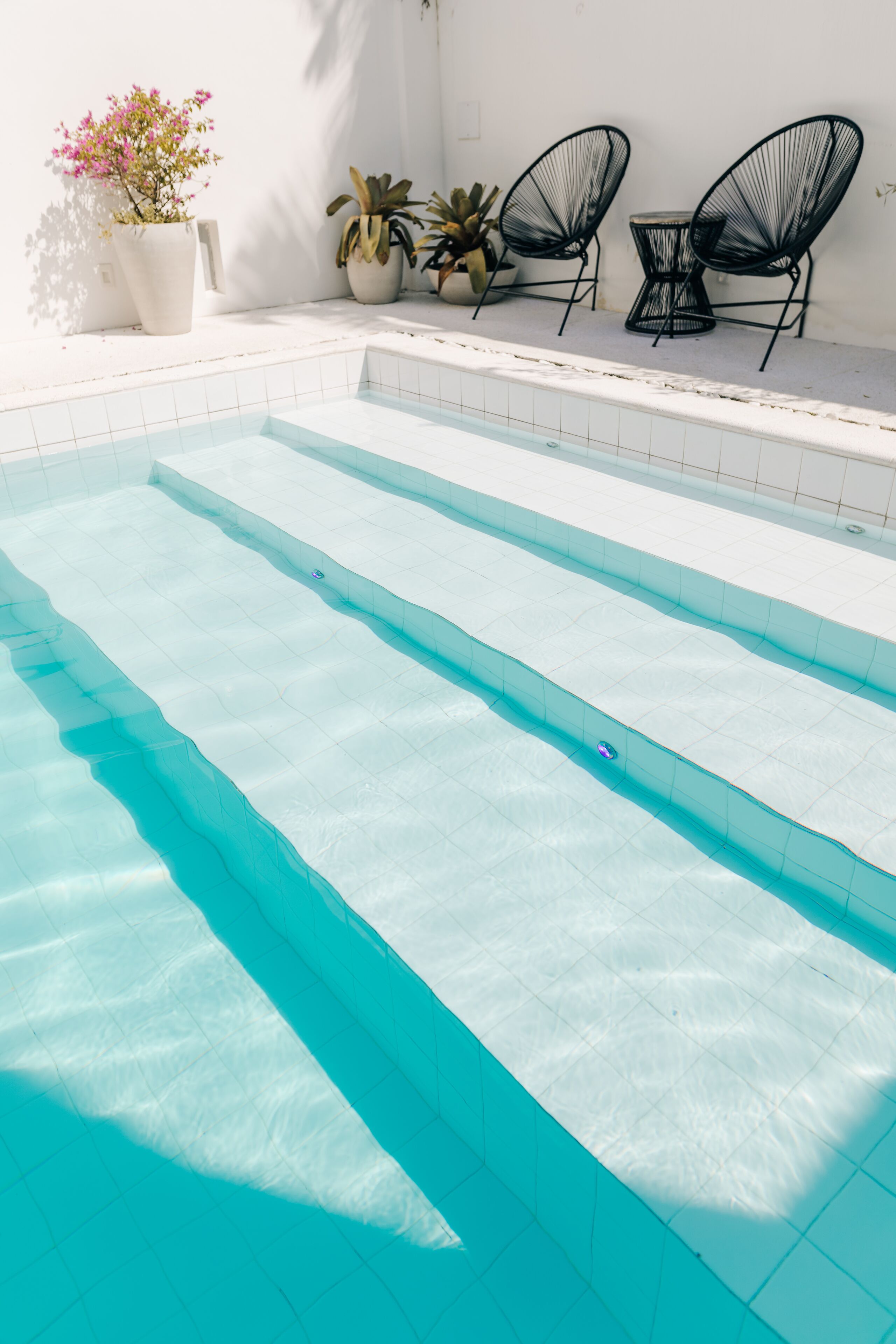 Uma piscina com água azul cristalina, rodeada por azulejos brancos. Há plantas em vasos e duas cadeiras de arame preto à beira da piscina.