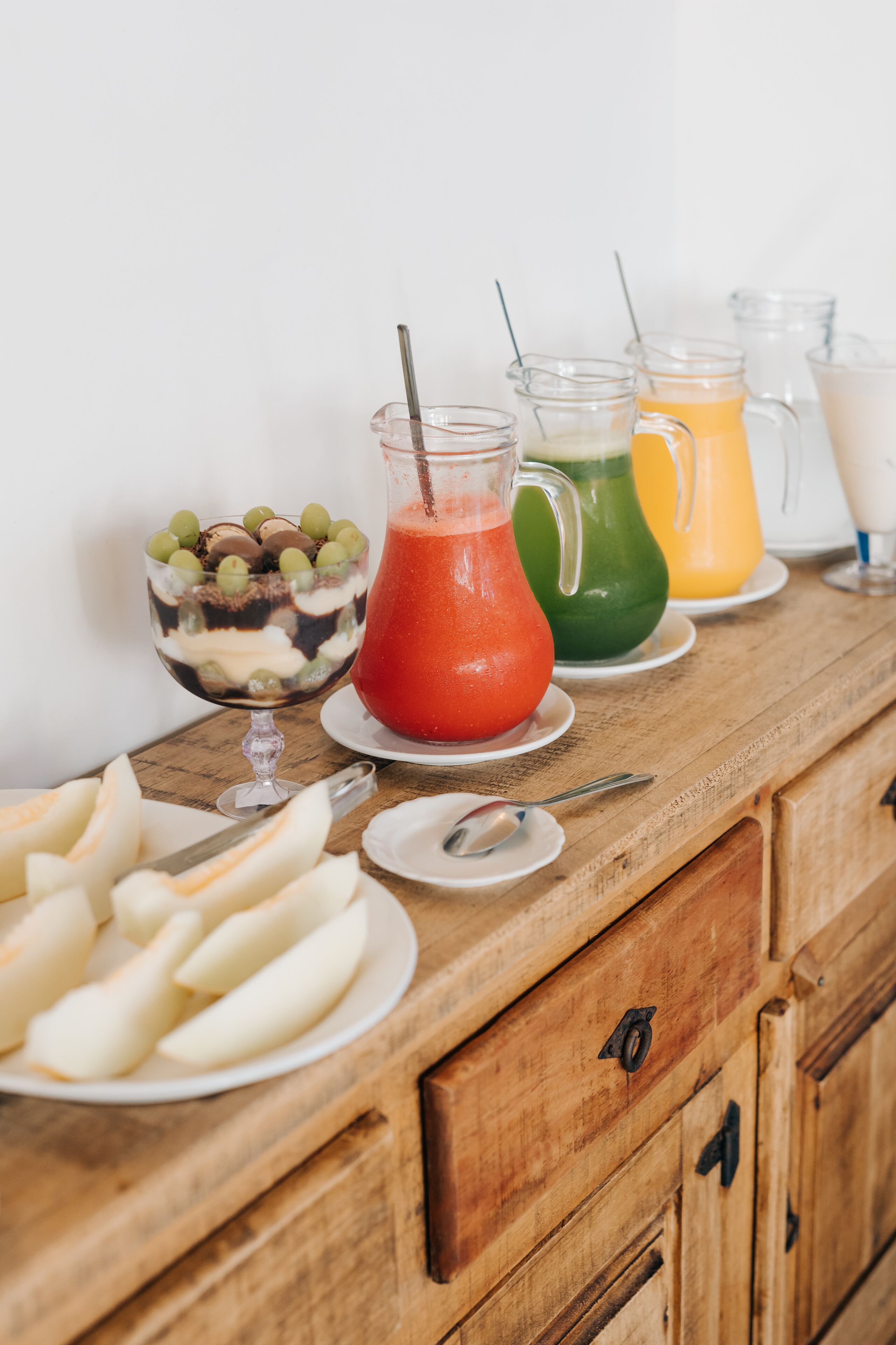 A wooden buffet table with a variety of food items and drinks, including sliced fruits, a layered dessert, and colorful beverages in pitchers.