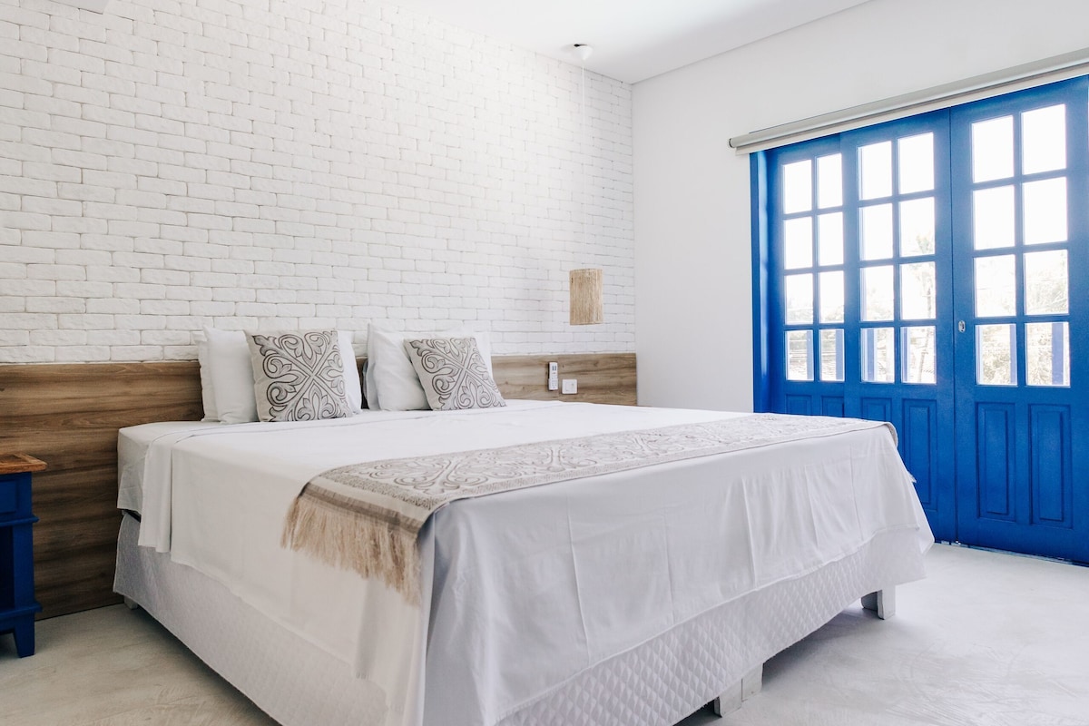 A bedroom with a white brick wall, a wooden headboard, a white bed with patterned pillows, a blue door, and a small bedside table.