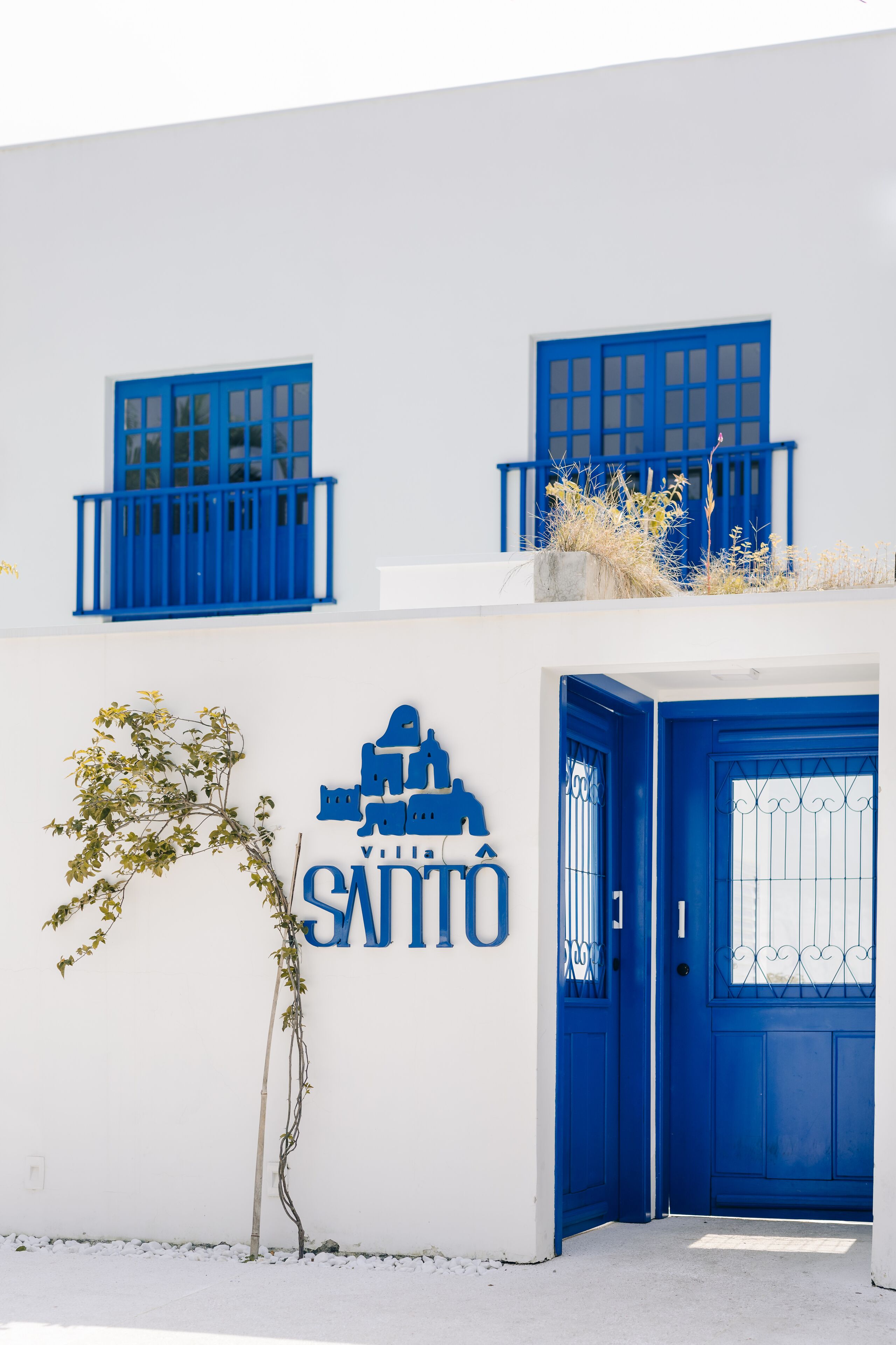 A building with a white facade and blue accents, including a blue door and windows. A sign with the name "Villa SANTOS" is visible.
