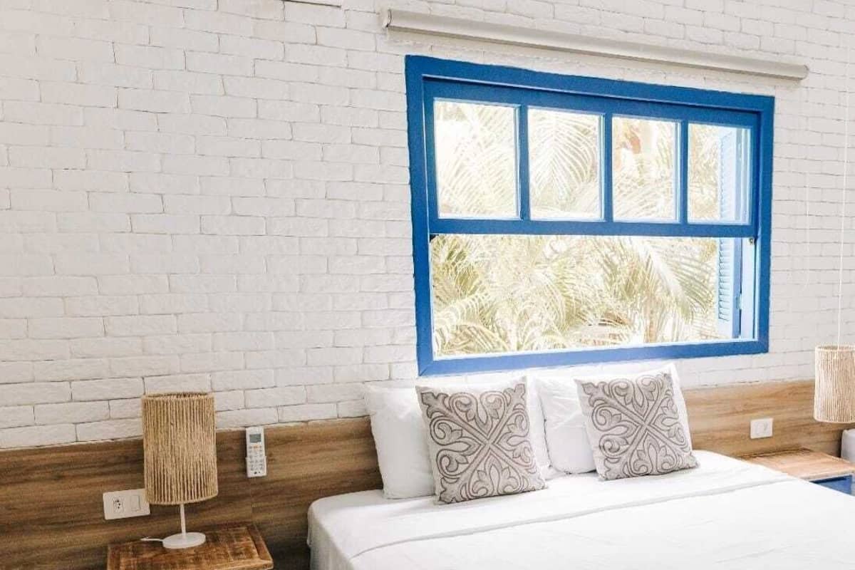 A bedroom with a white brick wall, a wooden headboard, a large window with blue trim, a bed with white bedding, two bedside tables with lamps, and a patterned throw on the bed.