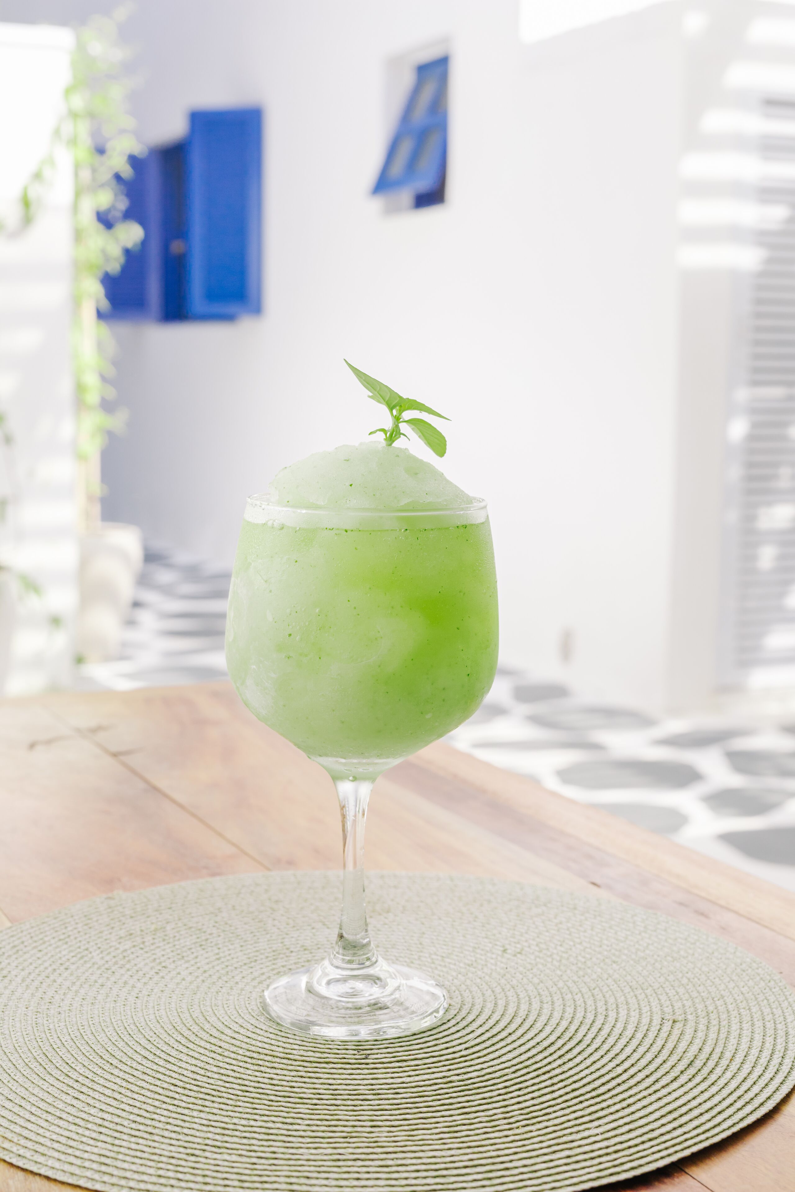 A green cocktail in a glass on a woven mat, with a white wall and blue shutters in the background.