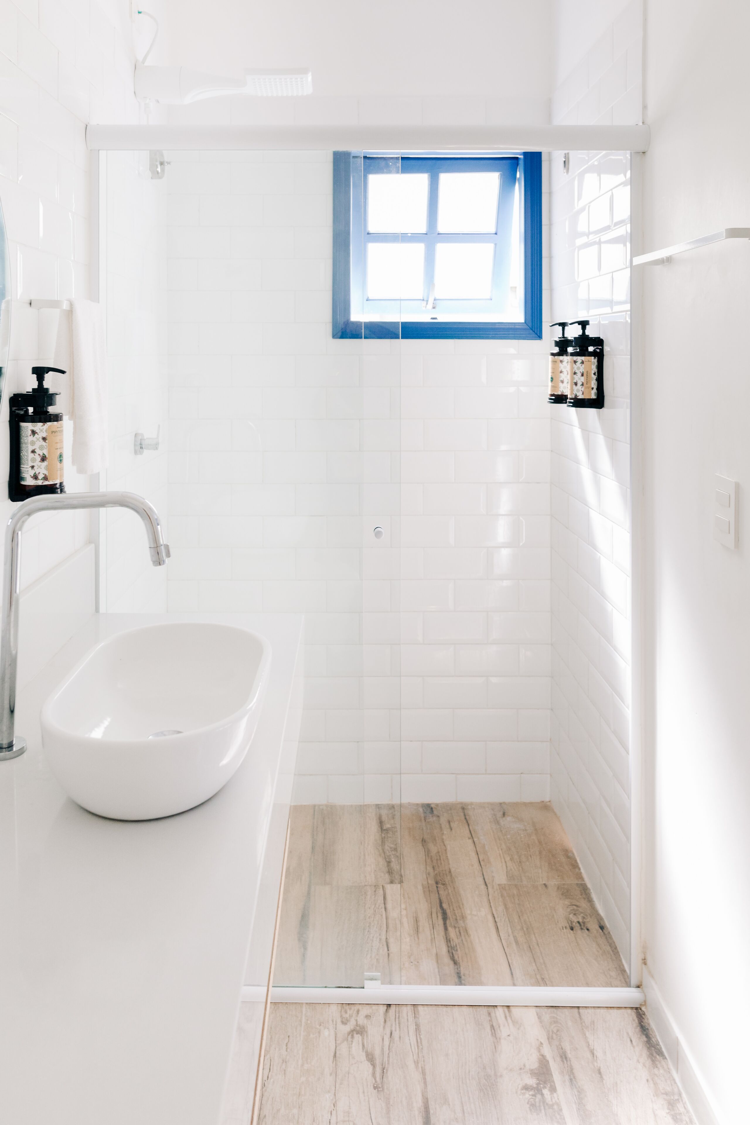 Un baño con lavamanos blanco, ducha con piso de madera y una ventana pequeña con detalles azules.