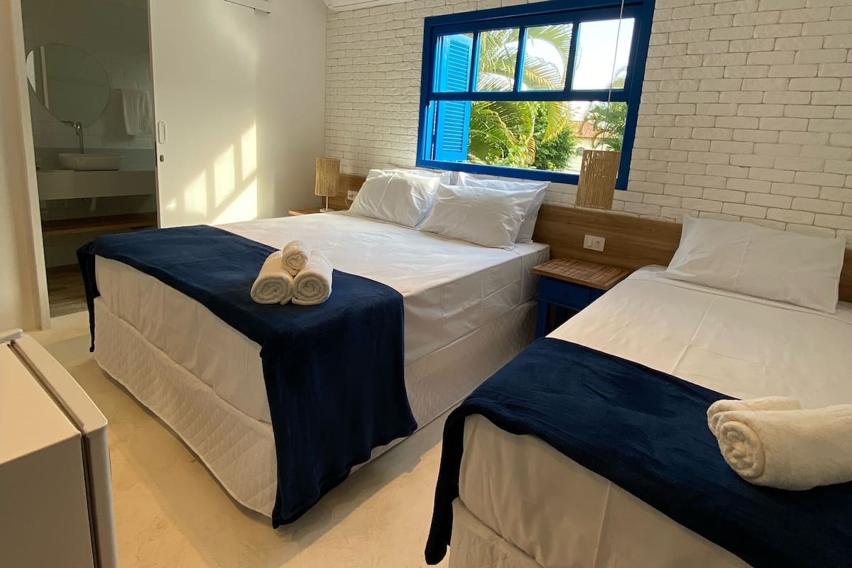 A room with two beds, a window with blue shutters, a white brick wall, and an air conditioning unit.
