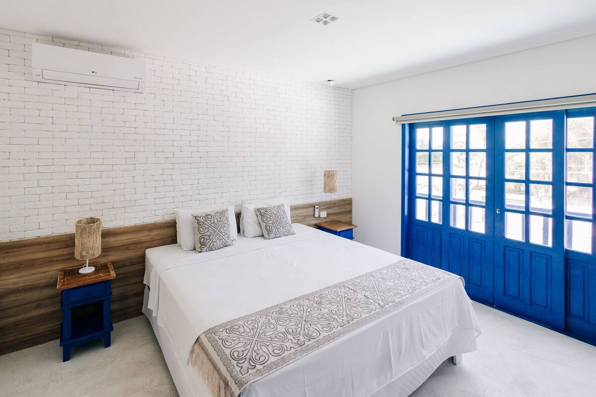 A bedroom with a white brick wall, a wooden headboard, a large bed with a white bedspread, two bedside tables with lamps, and a blue door with windows.