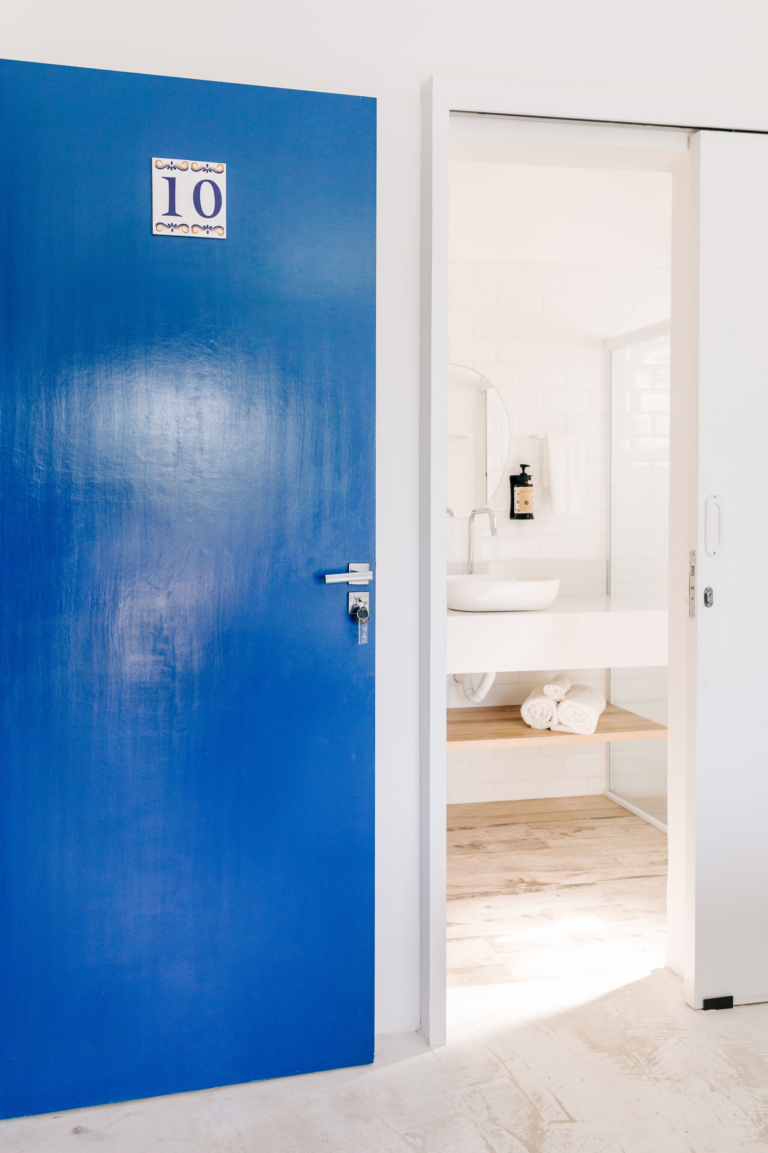Een kamer met een blauwe deur waarop '10' staat, een witte wastafel, een spiegel en een houten plank met handdoeken.