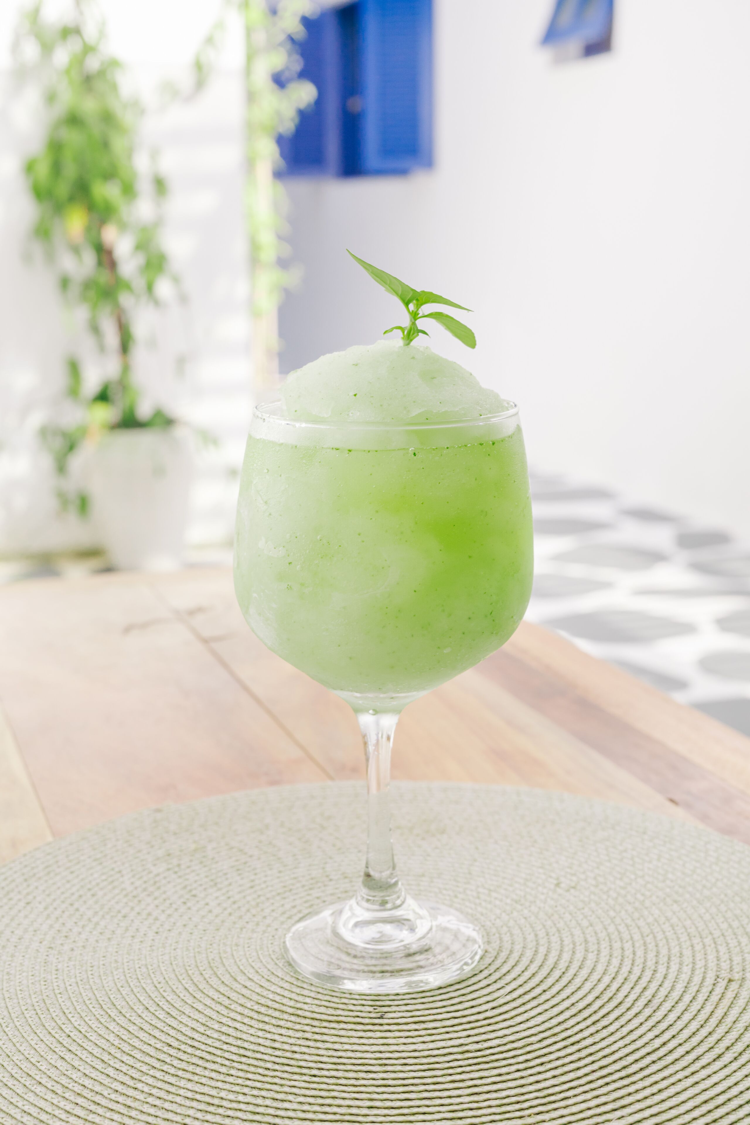 A green cocktail in a glass with a sprig of rosemary, placed on a woven mat.