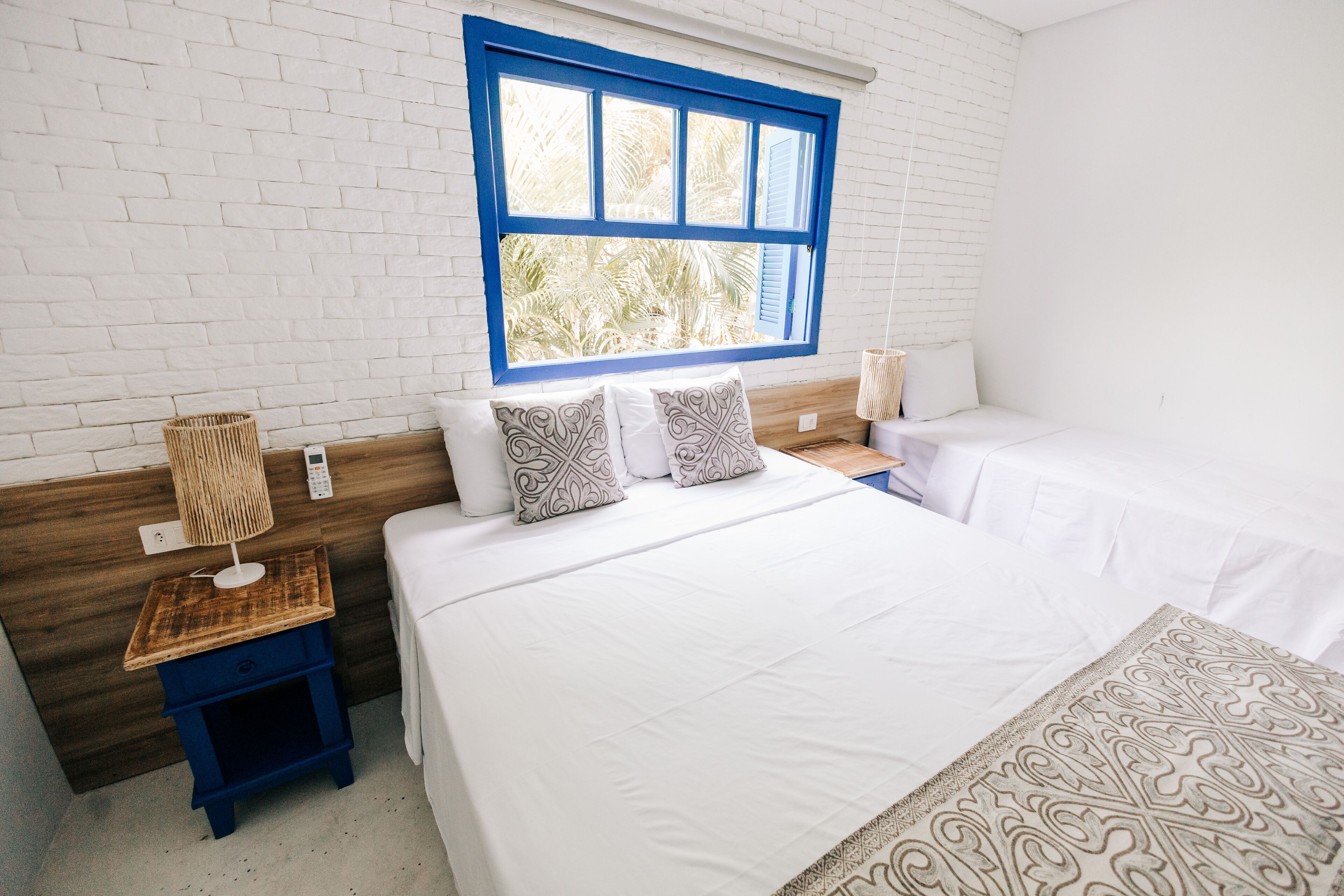 Un dormitorio con pared de ladrillo blanco, un ventanal con detalles azules, dos camas, una mesita de noche con lámpara y un edredón estampado sobre la cama.