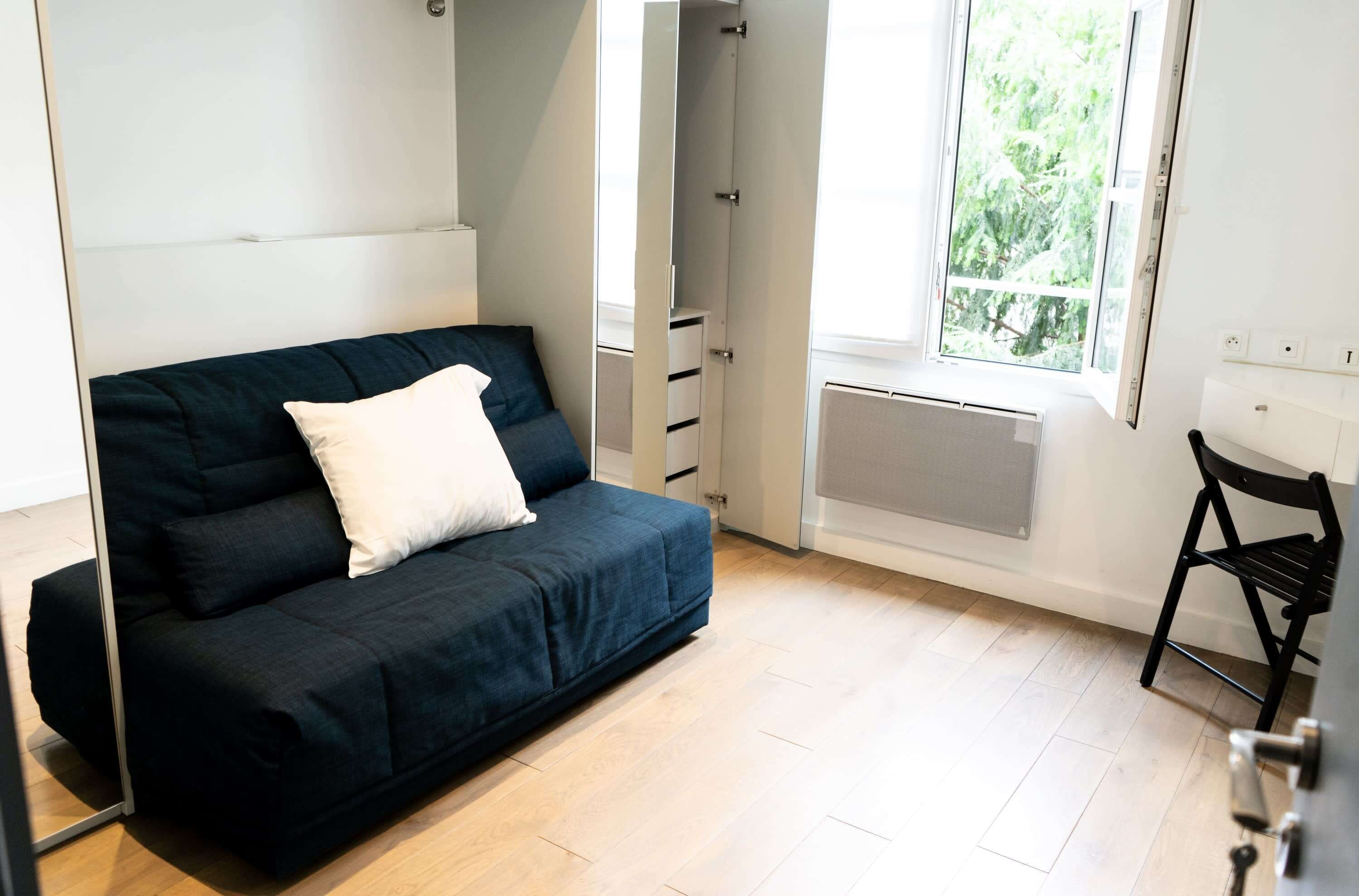 A modern, compact room with a dark blue sofa, a white pillow, a black chair, and a window with a view of greenery.