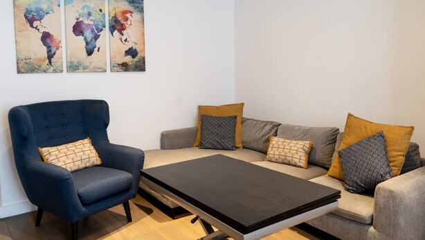 Ein modernes Wohnzimmer mit einem blauen Sessel, einem grauen Sofa mit gemusterten Kissen, einem schwarzen Couchtisch und abstrakten Weltkarten-Gemälden an der Wand.