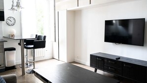 Een moderne woonkamer met een flatscreen televisie, een eethoek met een zwarte tafel en stoelen, en een keuken met een klok en hanglampen.