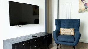 Een moderne woonkamer met een flatscreen televisie aan de muur, een donker houten kast met laden, en een blauwe fauteuil met een gestreept kussen.