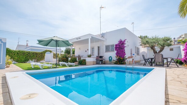 Une piscine rectangulaire bordée d’une terrasse blanche, un espace patio aménagé avec du mobilier d’extérieur, et un bâtiment blanc surmonté d’une antenne parabolique.