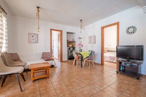 Un salon comprenant un canapé, des fauteuils, une table à manger, un téléviseur et un coin cuisine visible à travers une porte ouverte.
