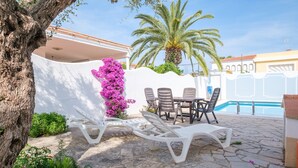 Un espace aménagé au bord de la piscine, avec des chaises longues, une table et un mur blanc agrémenté d’une composition florale rose.