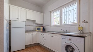 Une cuisine avec des meubles blancs, un four à micro-ondes, un lave-linge et une fenêtre avec des stores.