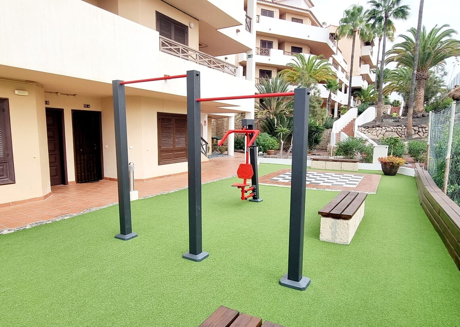 Un patio con césped artificial, equipo de ejercicio, una zona a cuadros y un edificio con balcones y puertas.