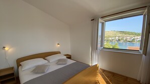 A bedroom with a wooden bed, two bedside tables, a nightstand, a window with a view of a lake and houses, and a wall-mounted lamp. - Apartments Pulic (Zaboric)
