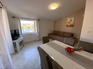A living room with a TV, a sofa, a dining table with a fruit bowl, and a window with a view of a body of water. - Apartments Pulic (Sibenik)