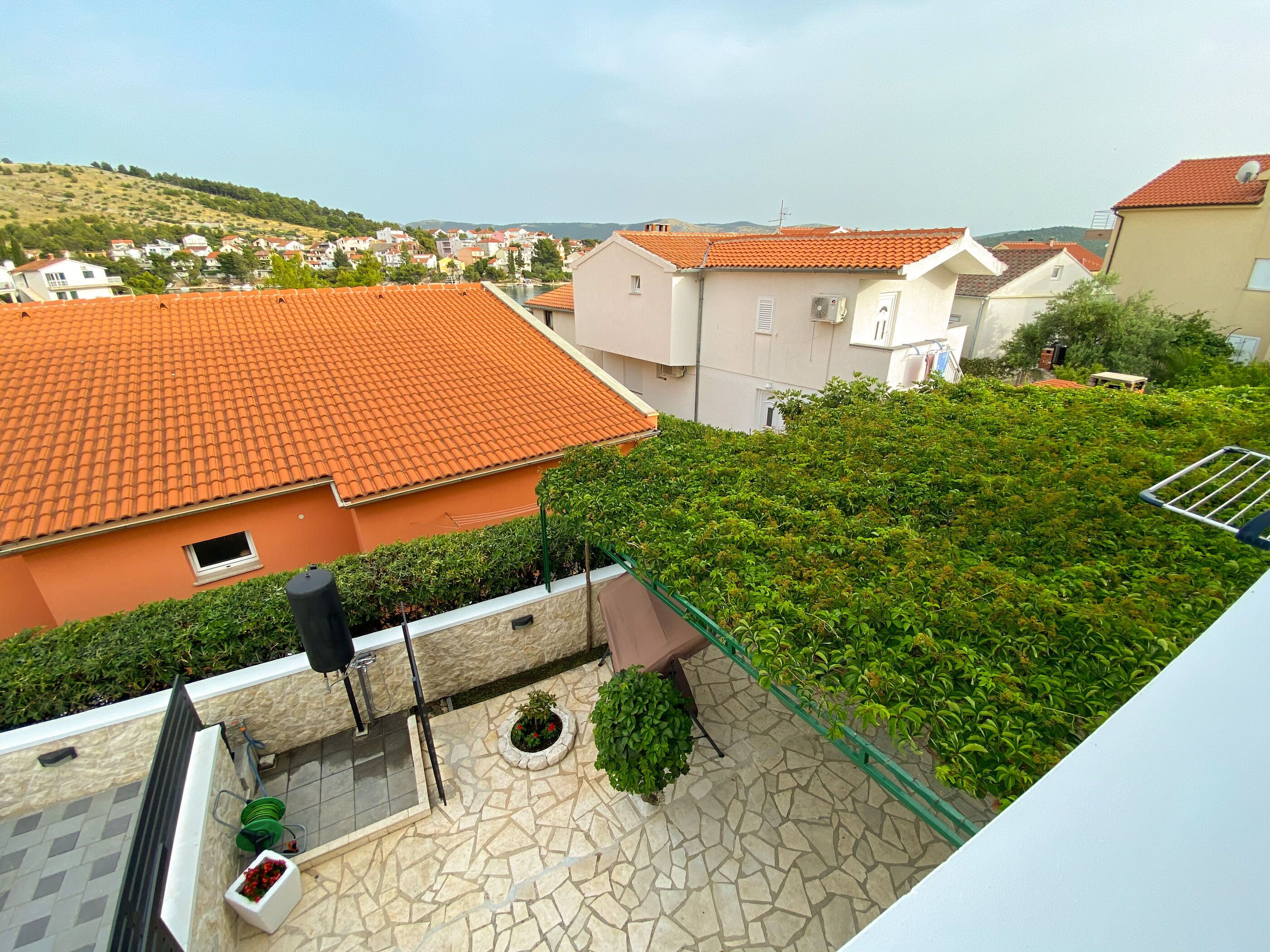 A rooftop terrace with a stone floor, green plants, and a view of a residential area with red-tiled roofs.