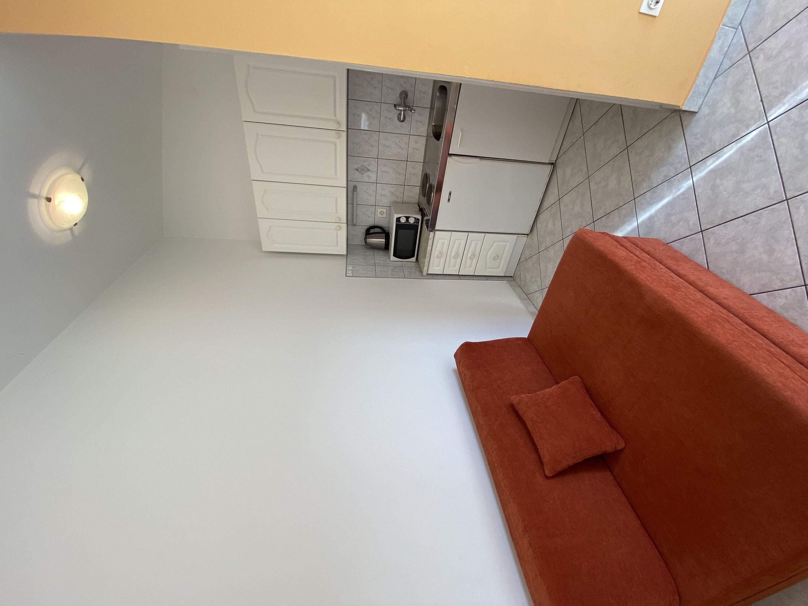Une pièce de taille modeste, avec un canapé blanc, un mur recouvert de carreaux et une petite cuisine équipée d’armoires et d’un four à micro-ondes.