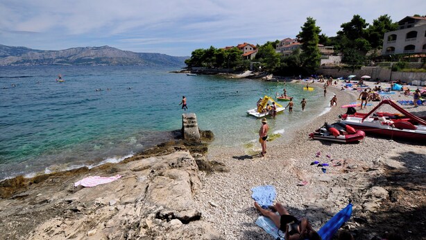 A beach with clear waters, rocky shore, and buildings in the background. - Apartments Postira uz More (Postira)
