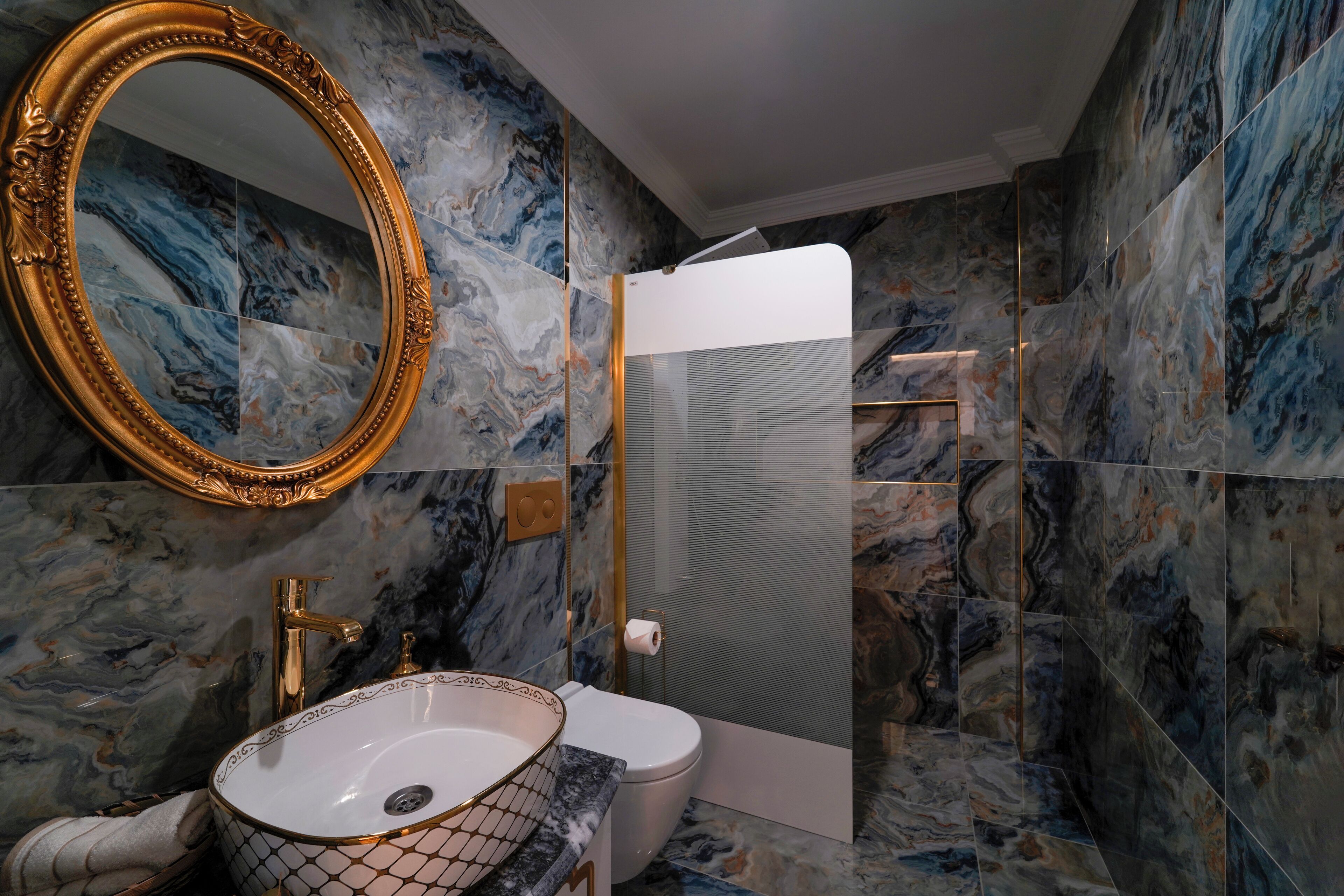 Un baño con pared de mármol, un espejo con marco dorado, un lavamanos blanco y una cabina de ducha de vidrio.