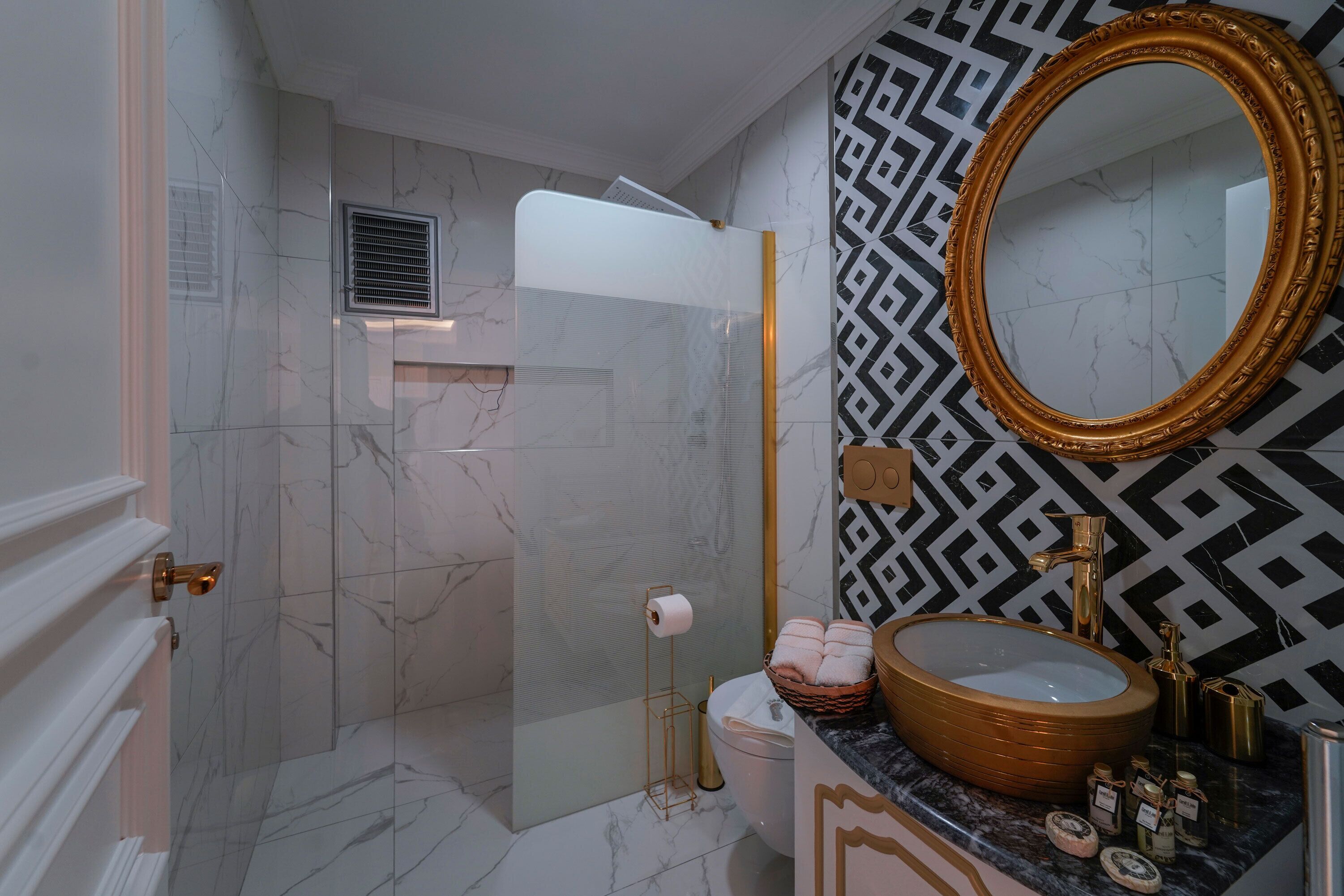 A bathroom with a marble floor, a large mirror with a gold frame, a sink with a gold basin, and a shower area.