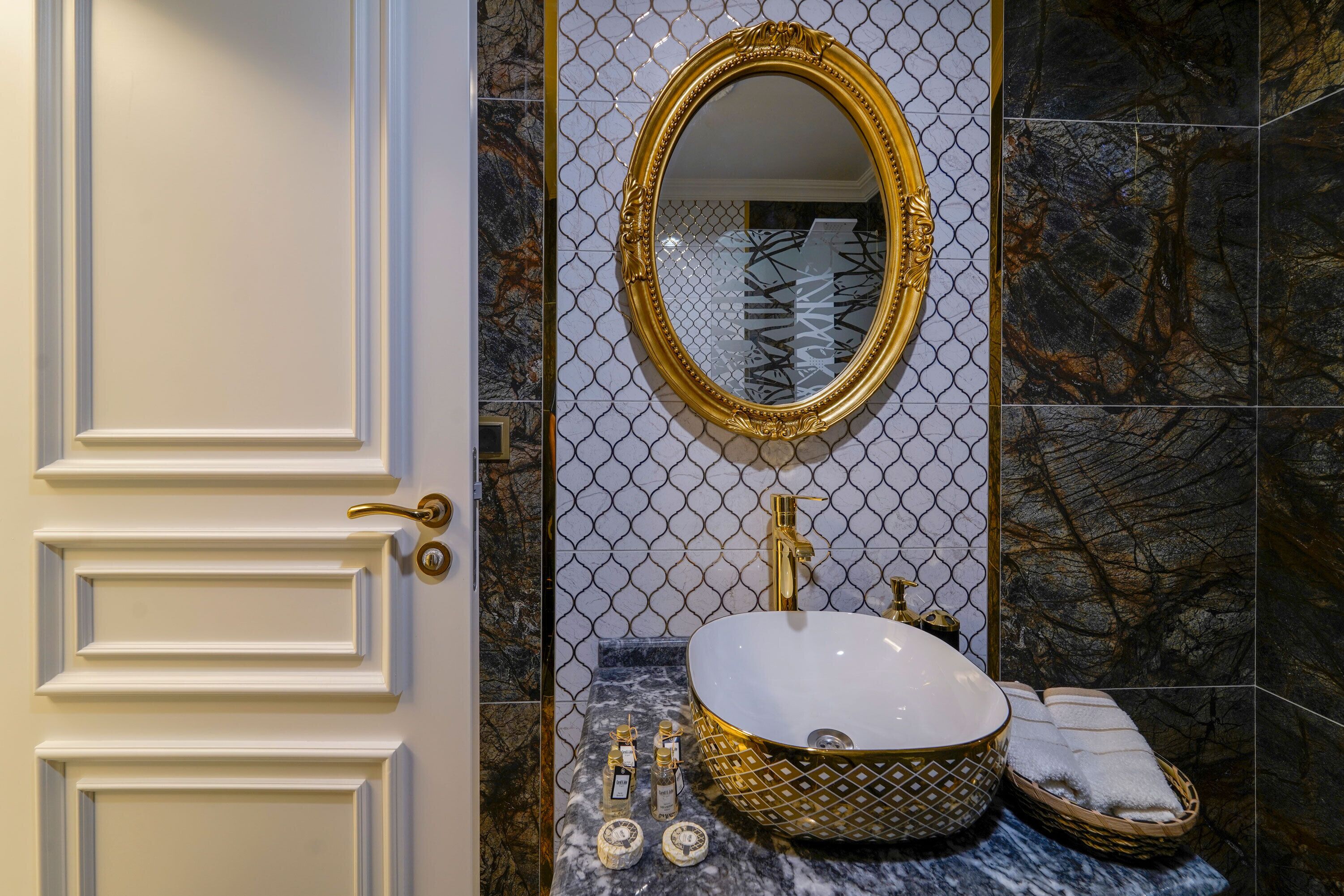 Un baño con puerta blanca, espejo con marco dorado, lavamanos de mármol y pared cubierta de azulejos con un diseño geométrico.