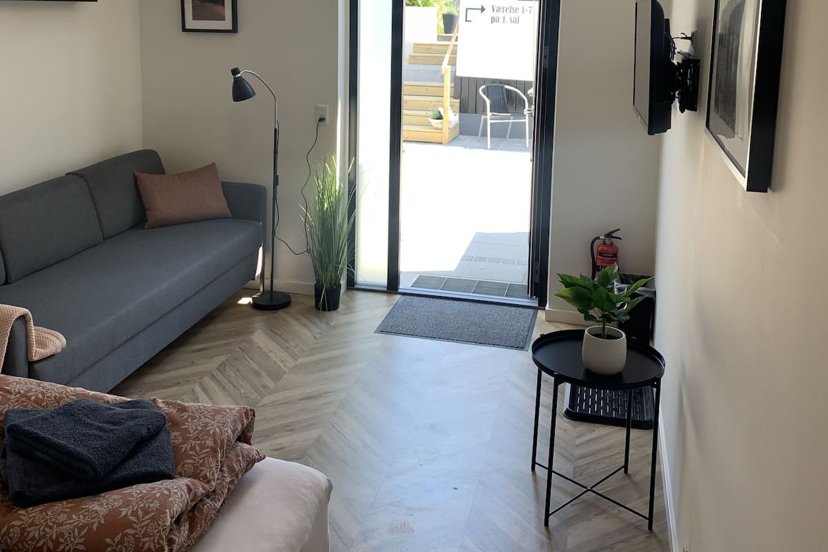 Un salon moderne avec un canapé gris, une petite table d’appoint agrémentée d’une plante, un lampadaire et une porte-fenêtre coulissante donnant sur un espace extérieur.