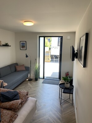 Una sala de estar moderna con un sofá gris, una mesita auxiliar con una planta, una lámpara de pie y un ventanal corredero que da a un espacio al aire libre.